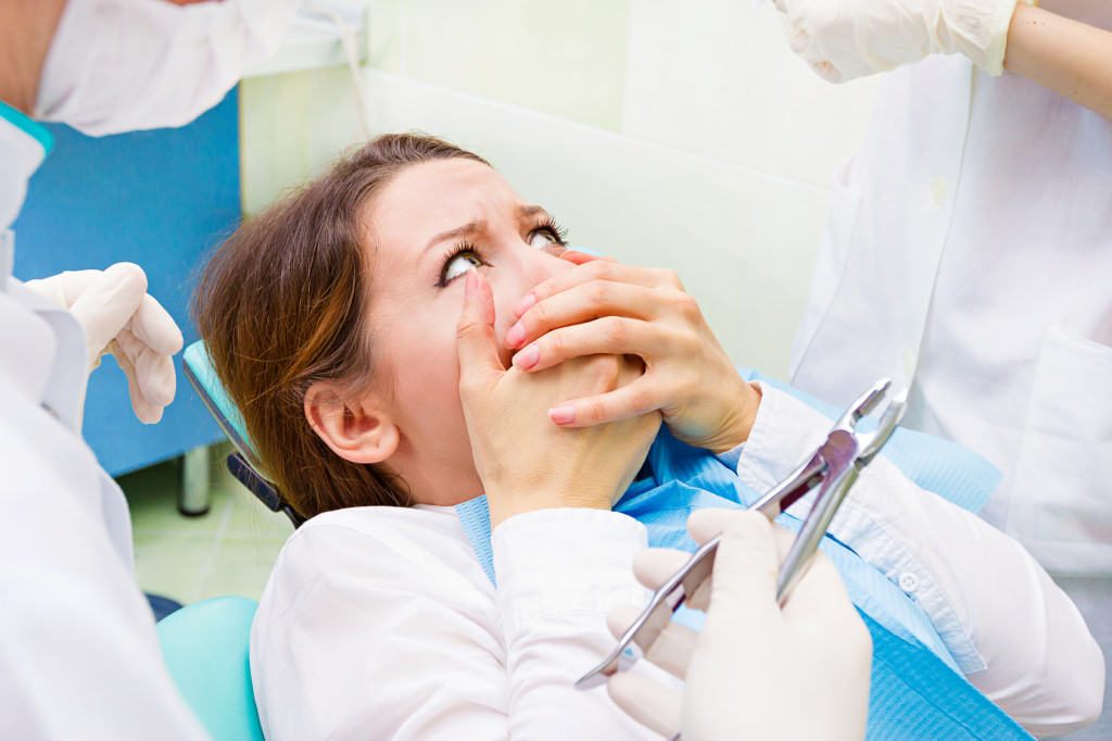 Closeup portrait young terrified girl woman scared at dentist visit, siting in chair, covering her mouth, doesn't want dental procedure, drilling, tooth extraction, isolated clinic office background